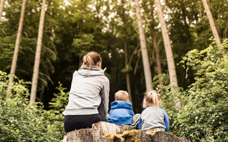 A mother and two young kids sit in a forest clearing
