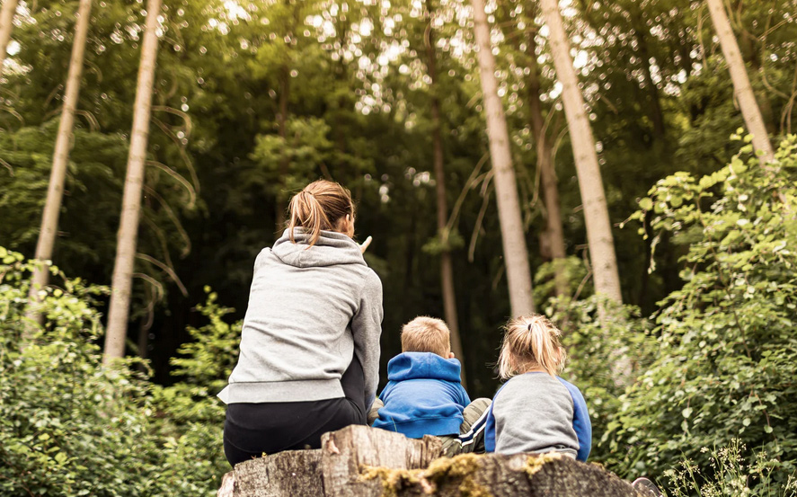 A mother and two young kids sit in a forest clearing
