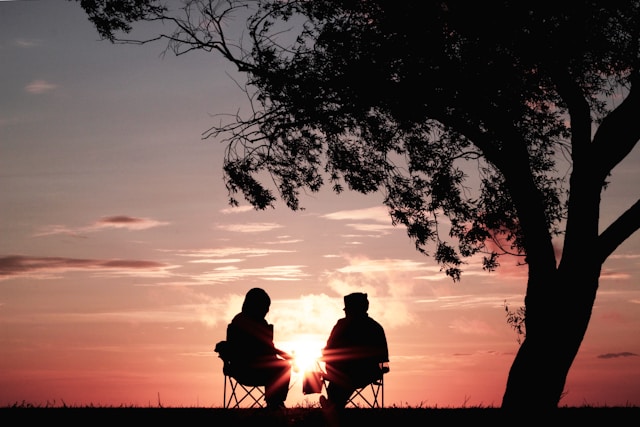 A couple sit under a tree in camp chairs with the morning sun behind them