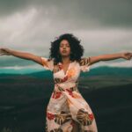 Woman of color in an dress holds her arms out with a dark sky behind her