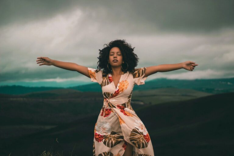Woman of color in an dress holds her arms out with a dark sky behind her